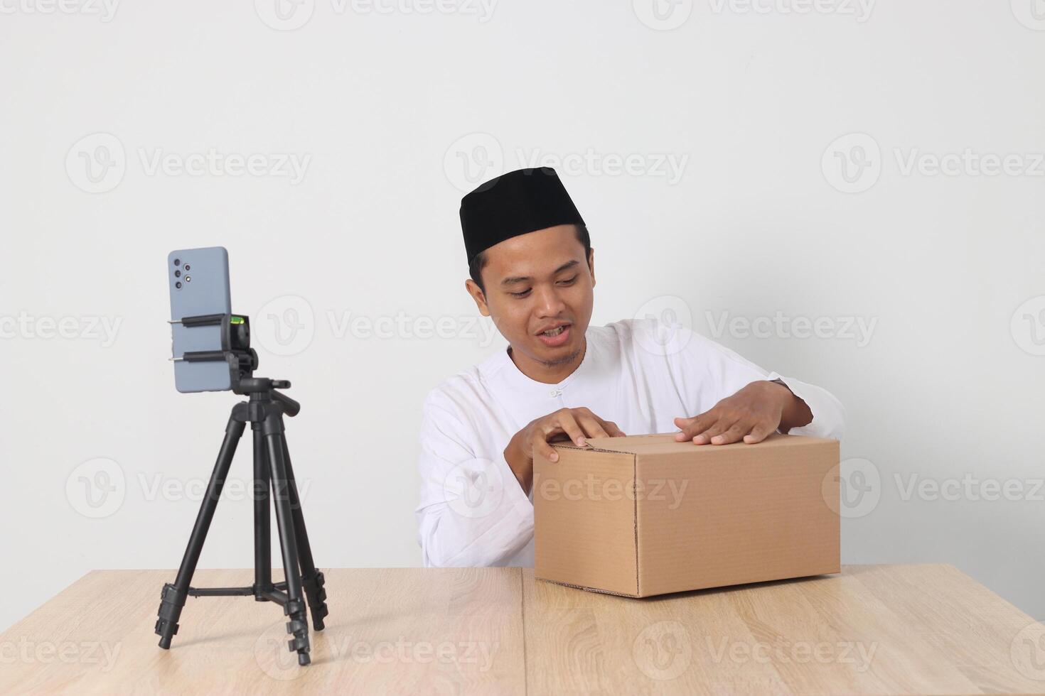 Portrait of excited Asian muslim man in koko shirt with skullcap promoting his product on live streaming session. Online shopping with smartphone concept. Isolated image on white background photo