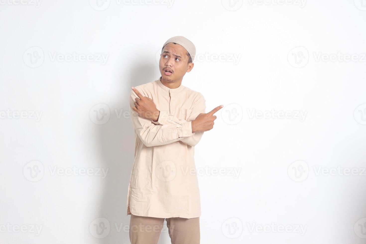 Portrait of excited Asian muslim man in koko shirt with skullcap pointing sideways, making choice, choosing between two objects, demonstrate product, crossed hands. Isolated image on white background photo
