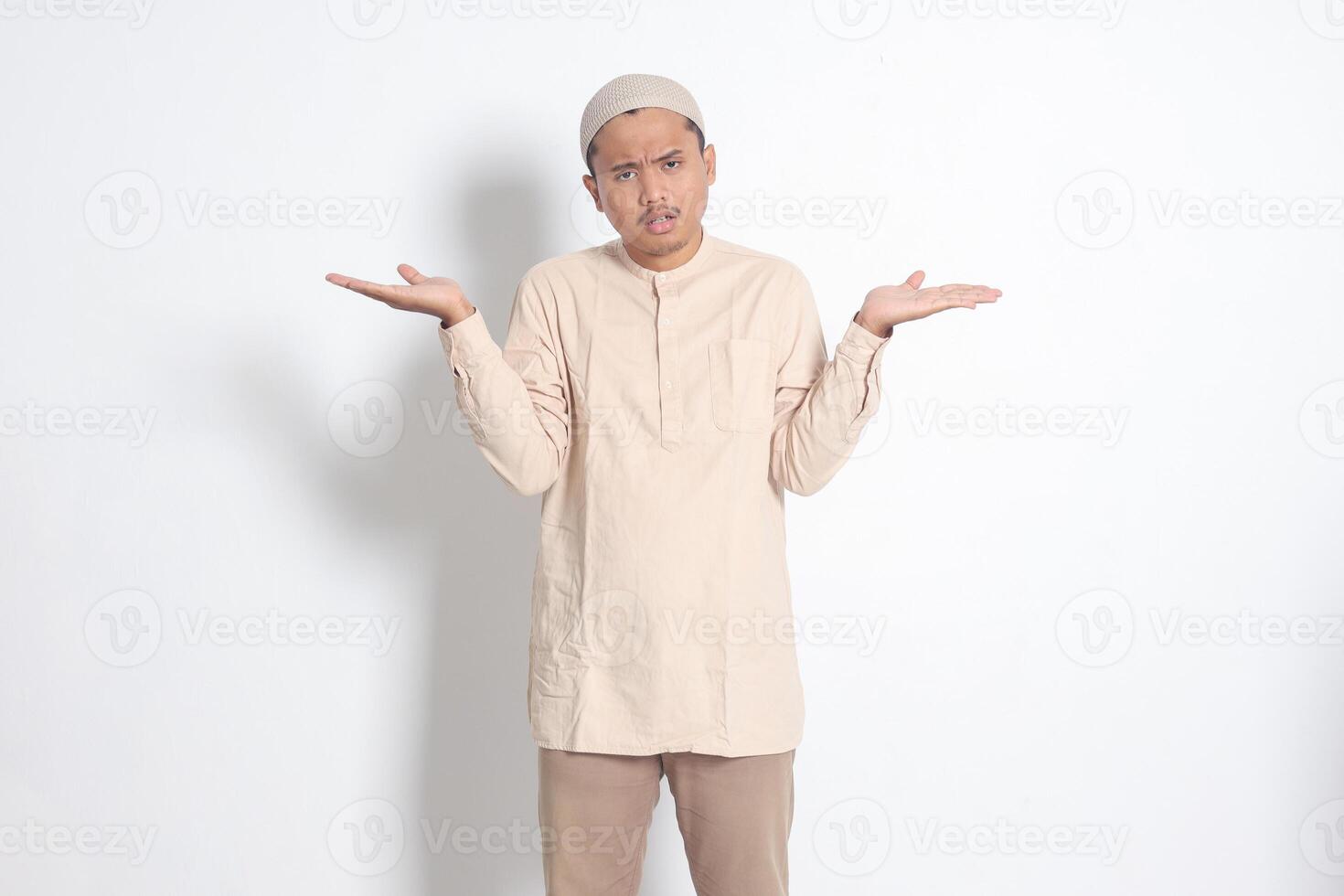 Portrait of confused Asian muslim man in koko shirt with skullcap spreading his hands sideways and holding two things, demonstrate products. Isolated image on white background photo
