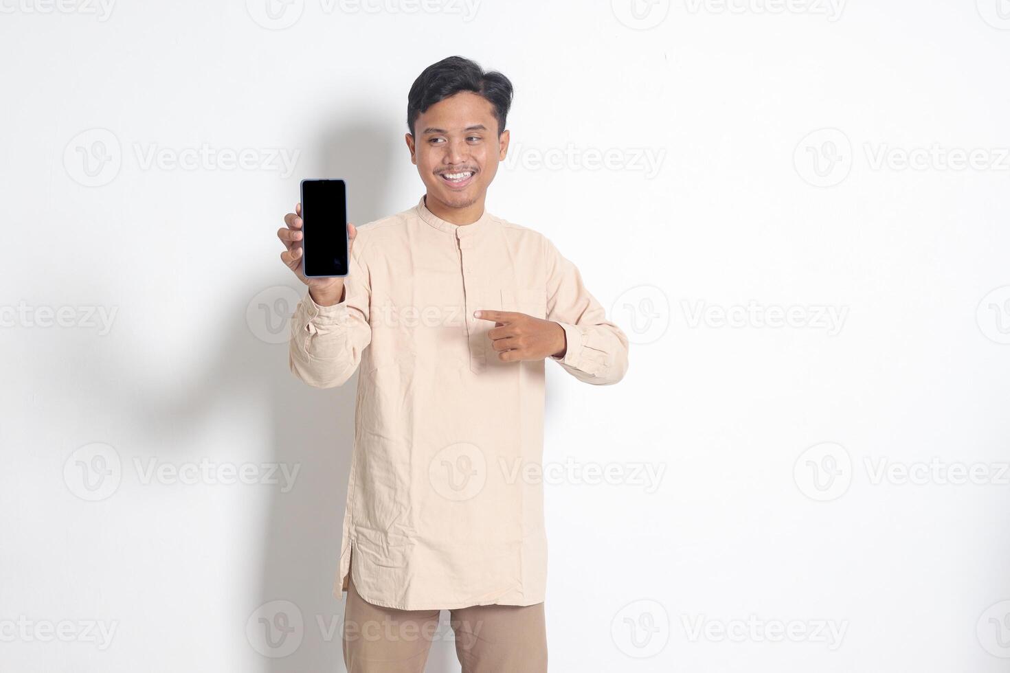 retrato de joven emocionado asiático musulmán hombre en koko camisa demostración blanco pantalla móvil teléfono Bosquejo mientras señalando y presentación producto. social medios de comunicación concepto. aislado imagen en blanco antecedentes foto