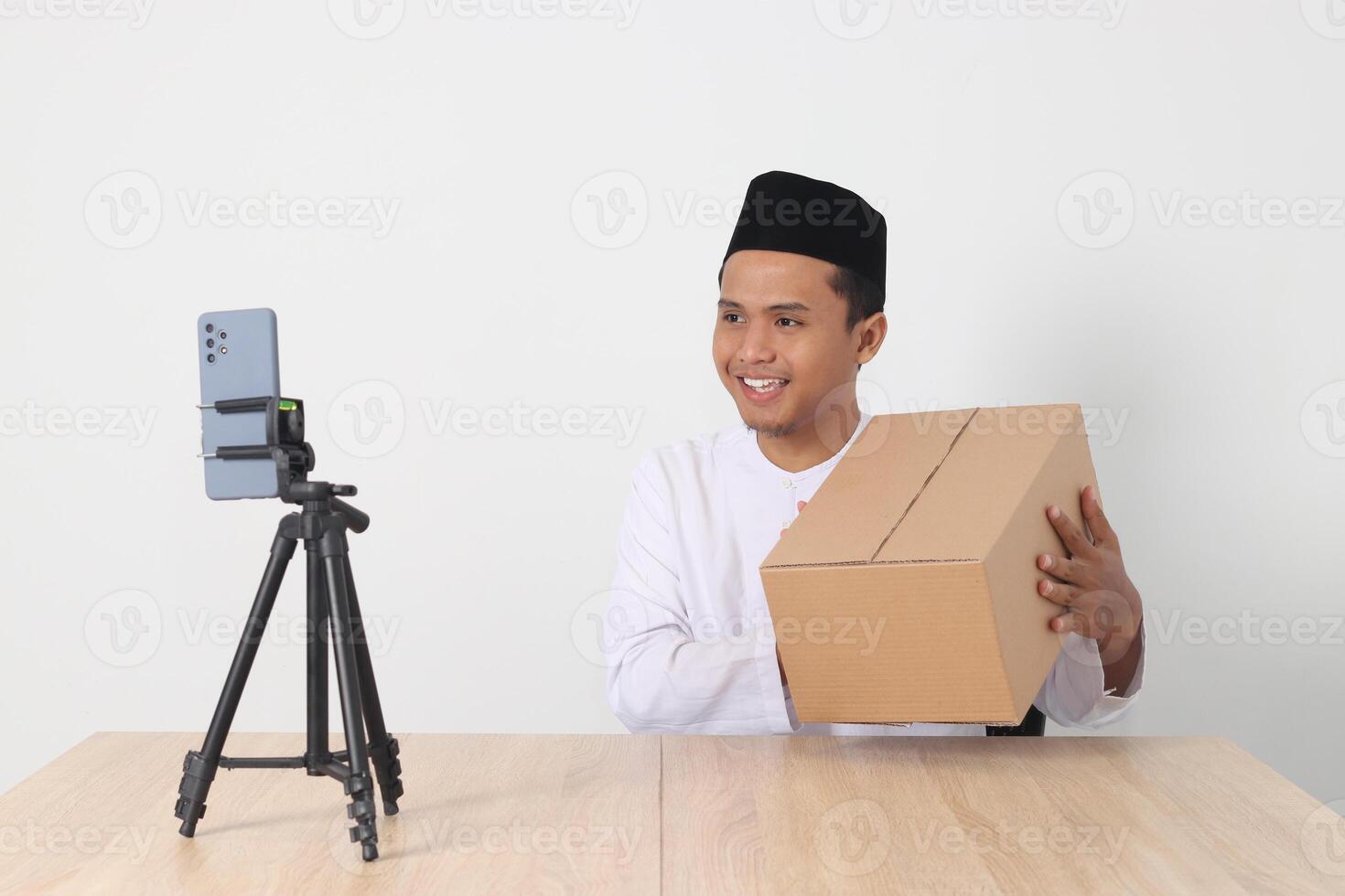 Portrait of excited Asian muslim man in koko shirt with skullcap promoting his product on live streaming session. Online shopping with smartphone concept. Isolated image on white background photo