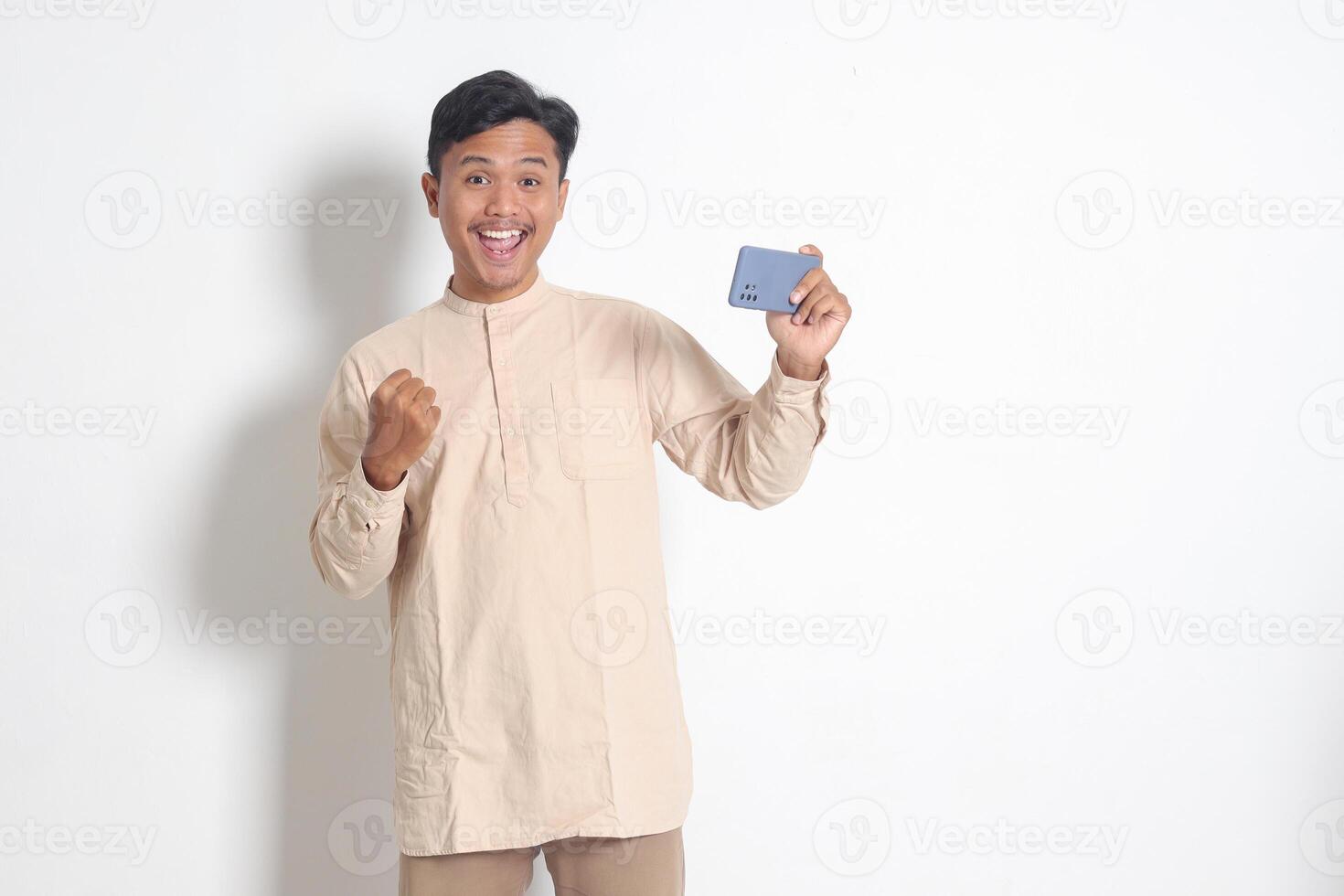 Portrait of excited Asian muslim man in koko shirt holding mobile phone and playing games on his smartphone, raising hands, celebrating success, become a winner. Isolated image on white background photo