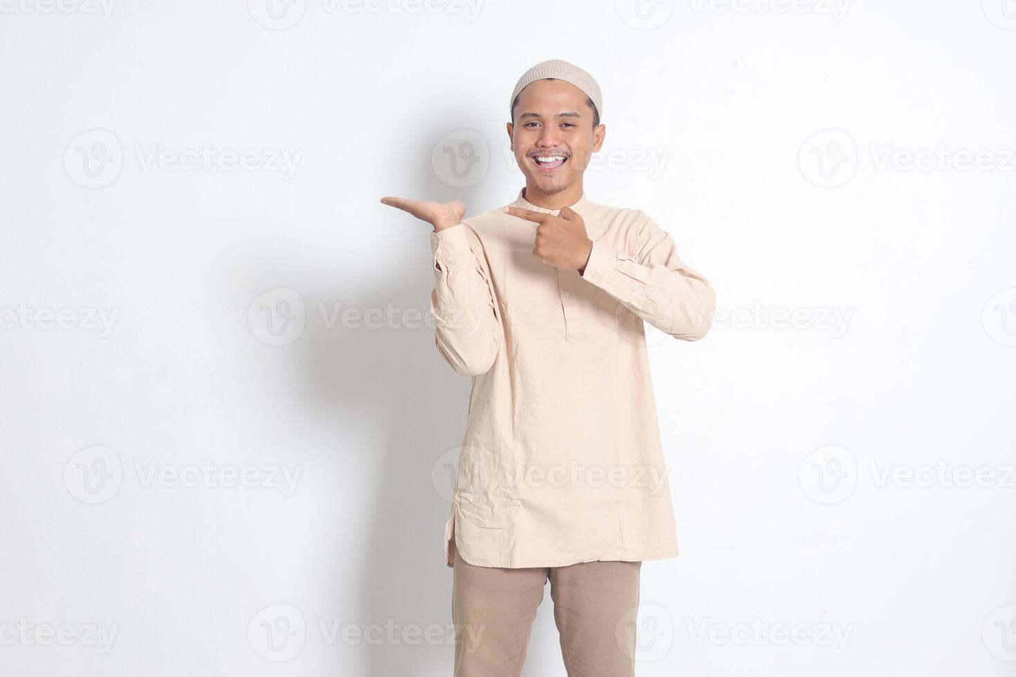 Portrait of shocked Asian muslim man in koko shirt with skullcap showing product and pointing with his hand and finger to the side. Advertising concept. Isolated image on white background photo