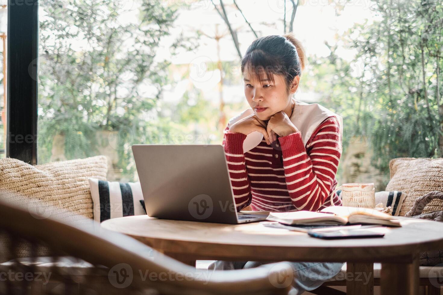 A focused woman sits absorbed in her work on a laptop at a round wooden table, surrounded by the comfort of a well-lit, plant filled room. photo