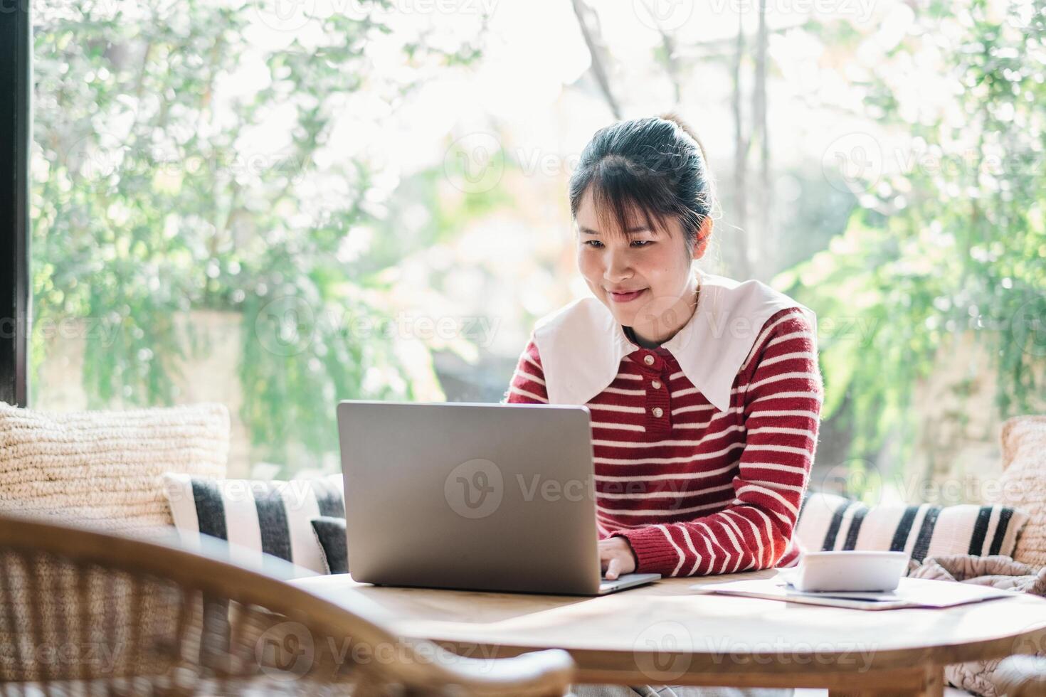 Cheerful woman enjoys a productive day working on her laptop in a well lit, plant-filled cafe, exuding a sense of satisfaction and comfort with a warm beverage by her side. photo