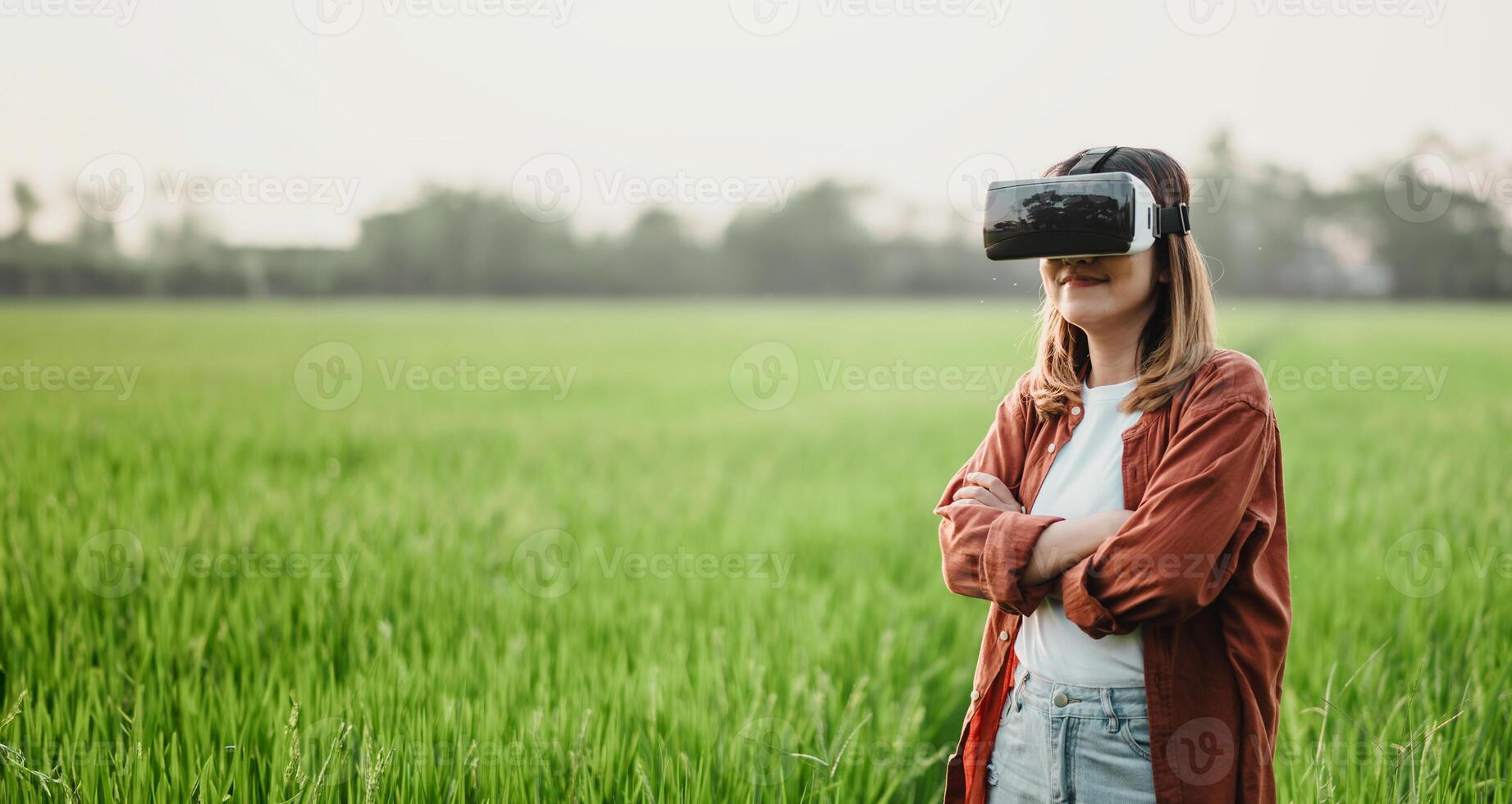 Young woman stands in a lush green field, arms crossed, immersed in a virtual reality world through her VR headset. photo