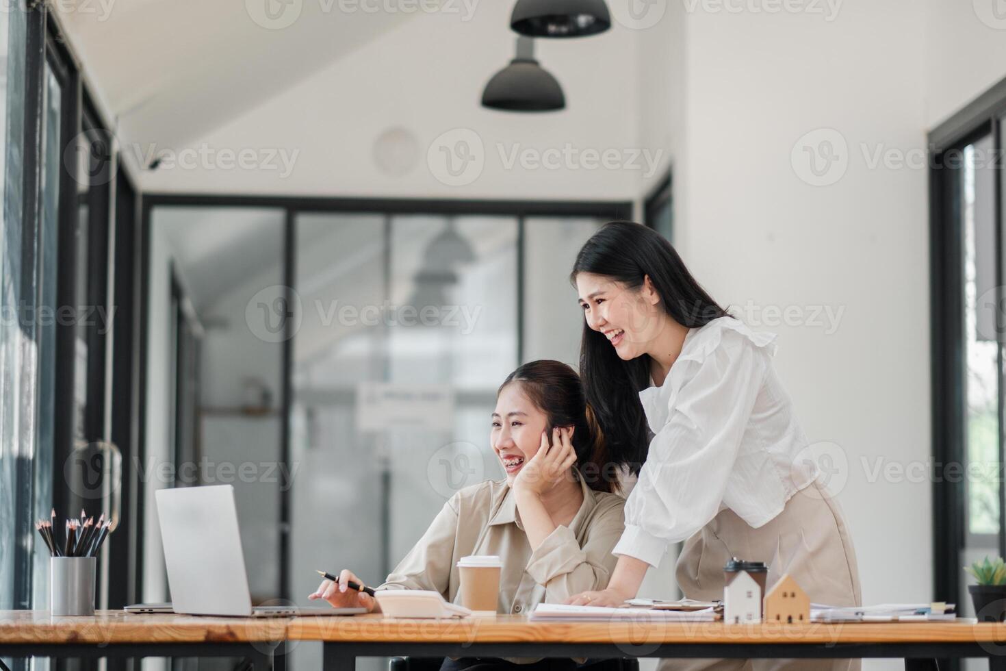 Two joyful colleagues share a light-hearted moment, with one seated at a laptop and the other standing beside her, in a spacious and well-lit office. photo