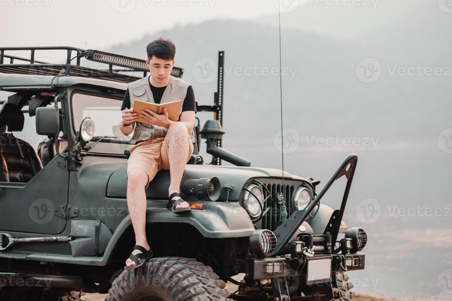 Young man sits thoughtfully on the hood of his car, reading a book, with mountains in the hazy distance. photo