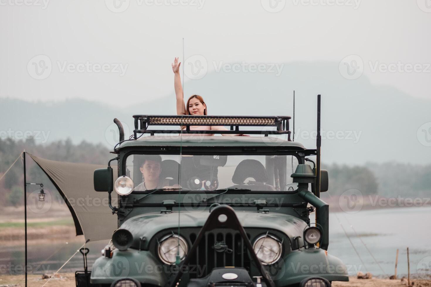 Woman on an adventure waves cheerfully from the roof of a off-road car parked by a tranquil lakeside campsite. photo