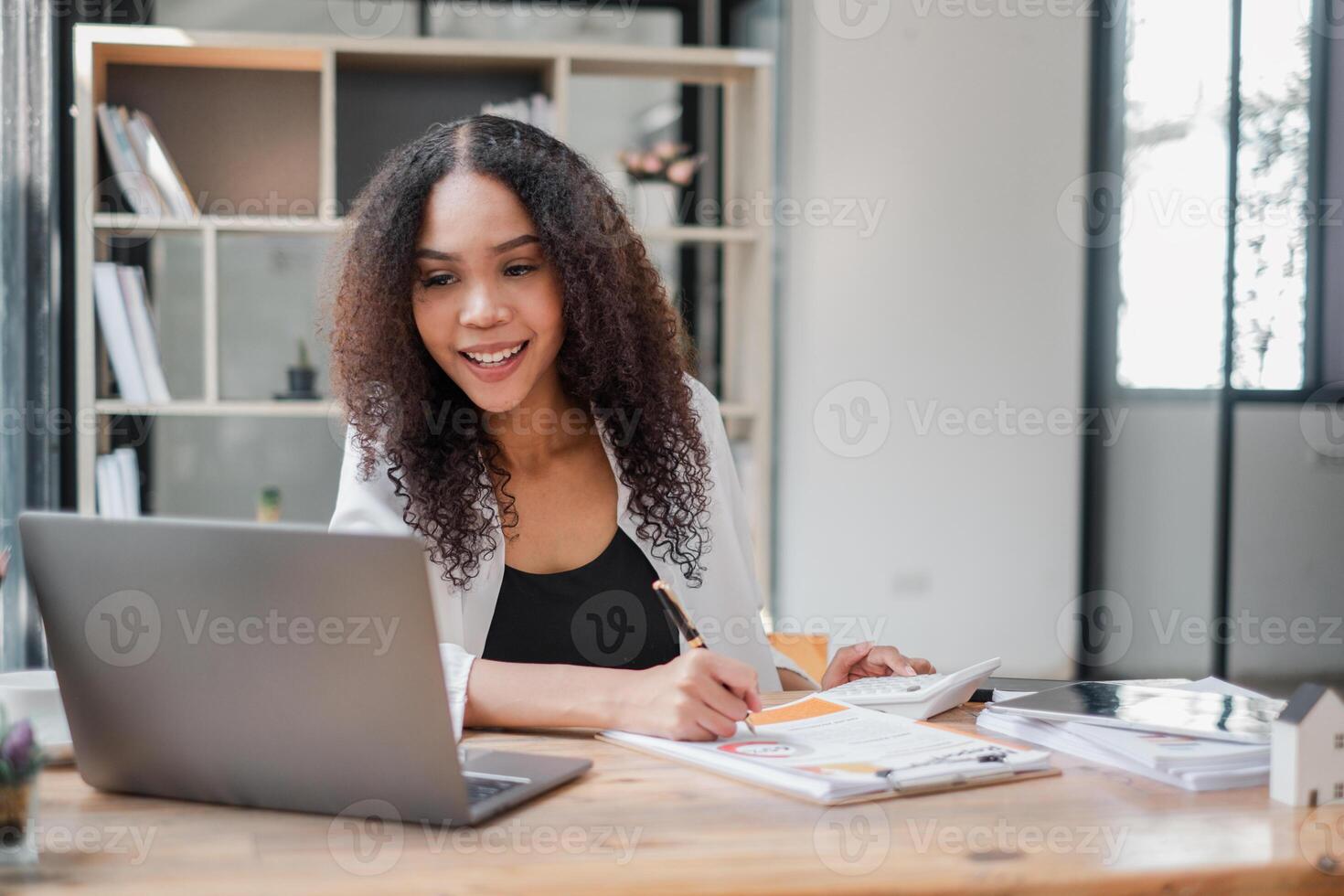 Smiling young businesswoman works on financial documents with a calculator and laptop at a sunny office desk. photo