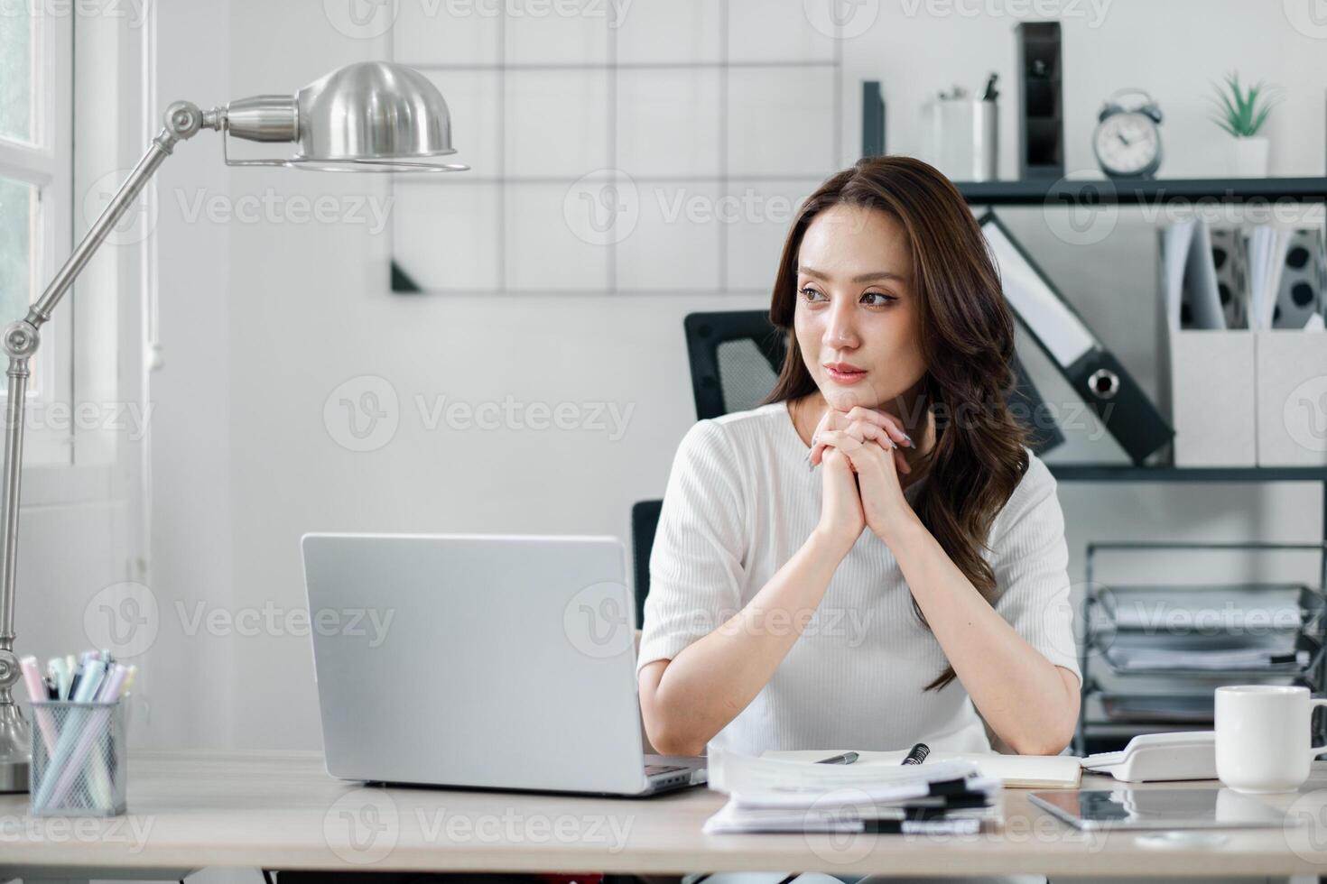 pensativo mujer de negocios con entrelazado manos se sienta a su limpiar y organizado escritorio en un oficina ajuste. foto