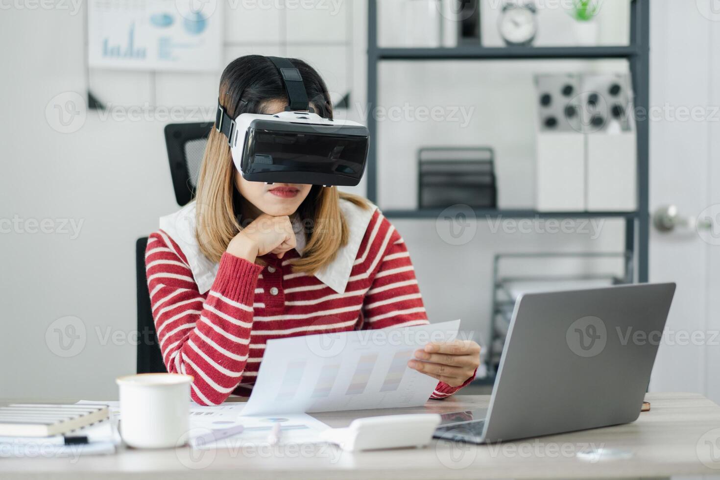 mujer en virtual realidad auriculares examina papel gráficos a un moderno trabajo escritorio con ordenador portátil y café. foto