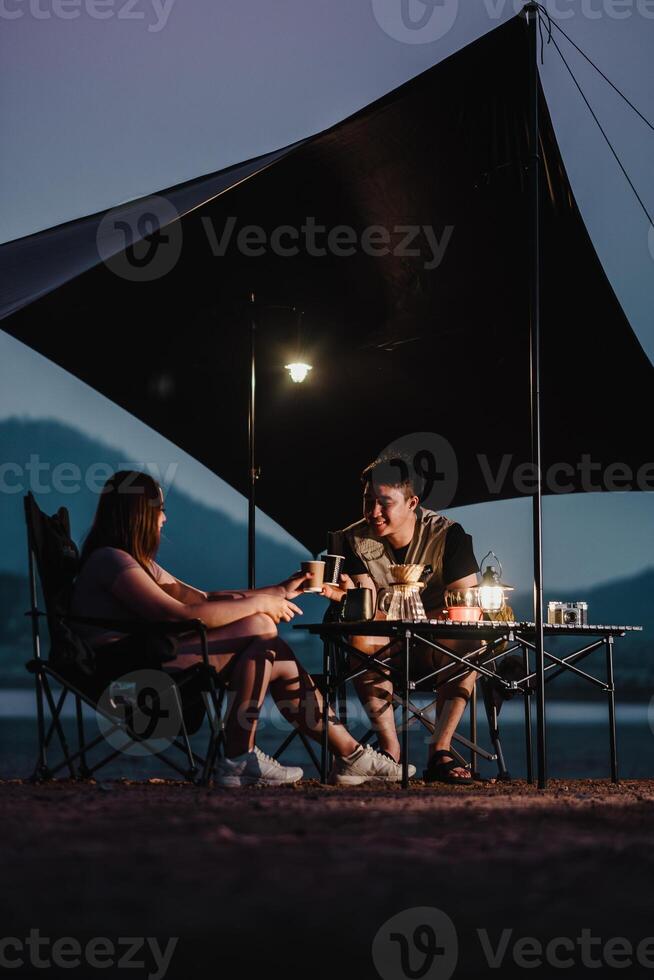 In the serene twilight, a couple shares a warm coffee at a well-equipped campsite, nestled by the lake under a spacious tarp. photo