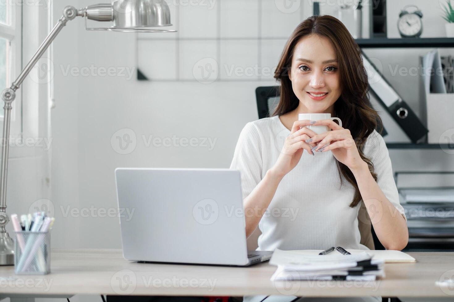 sonriente joven profesional mujer tomando un café descanso a su ordenado y elegante hogar oficina escritorio, encarnando un relajado trabajo atmósfera. foto
