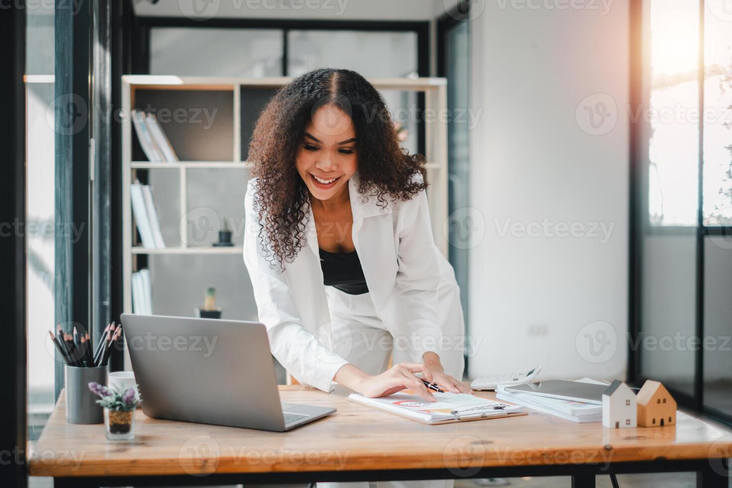 A radiant businesswoman leans over her work desk, thoroughly engrossed in organizing documents with a laptop nearby, in a sunlit modern office. photo