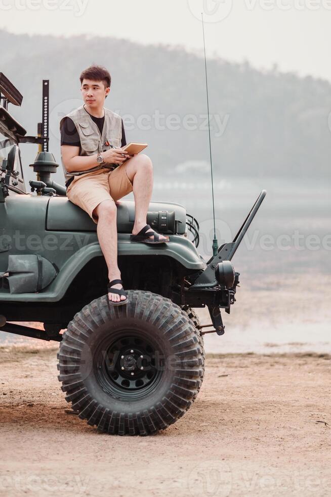 Seated on a large tire, a young man with a notebook looks contemplative against the serene backdrop of a lakeside landscape. photo