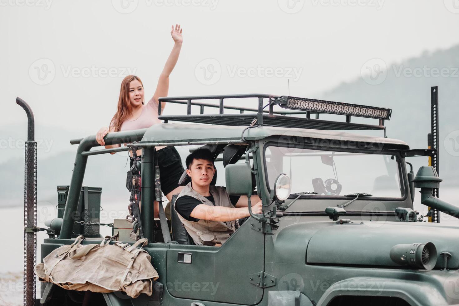 Woman waves cheerfully from the roof of a car while the man looks on, both ready for their next adventure on a road trip. photo