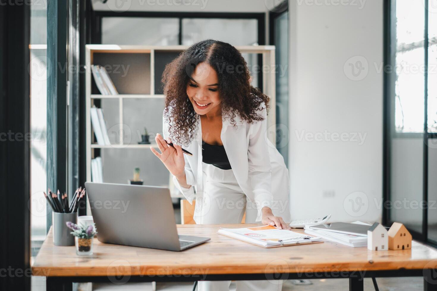 A charismatic businesswoman gestures during an animated conversation, likely on a video call, as she stands at her work desk in a bright, contemporary office space. photo