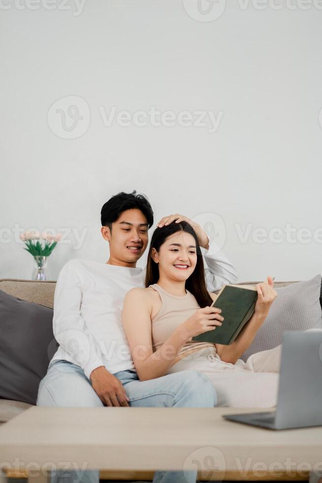 Young man and woman sit comfortably on a couch, reading a book together. The man, wearing a white shirt and jeans, has an arm around the woman as they both focus on the book. photo