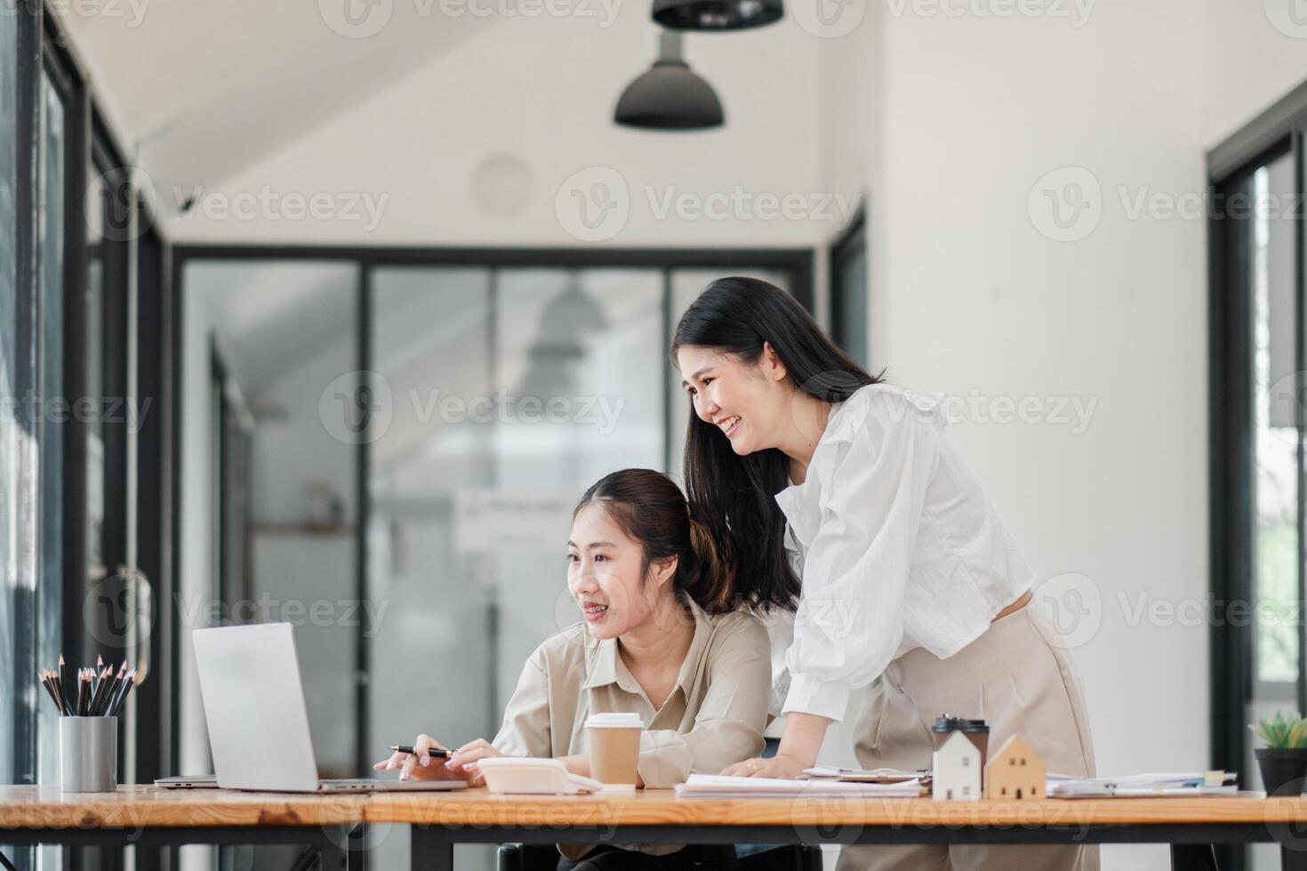 Two smiling female colleagues collaboratively working on a project with a laptop and coffee in a bright office space. photo