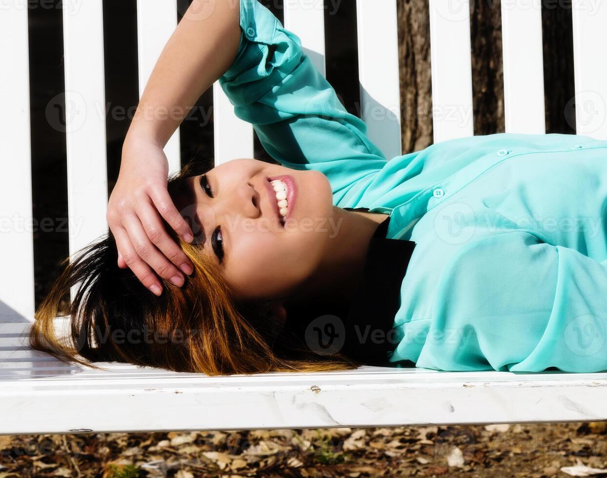 sonriente asiático americano mujer reclinable en banco al aire libre foto