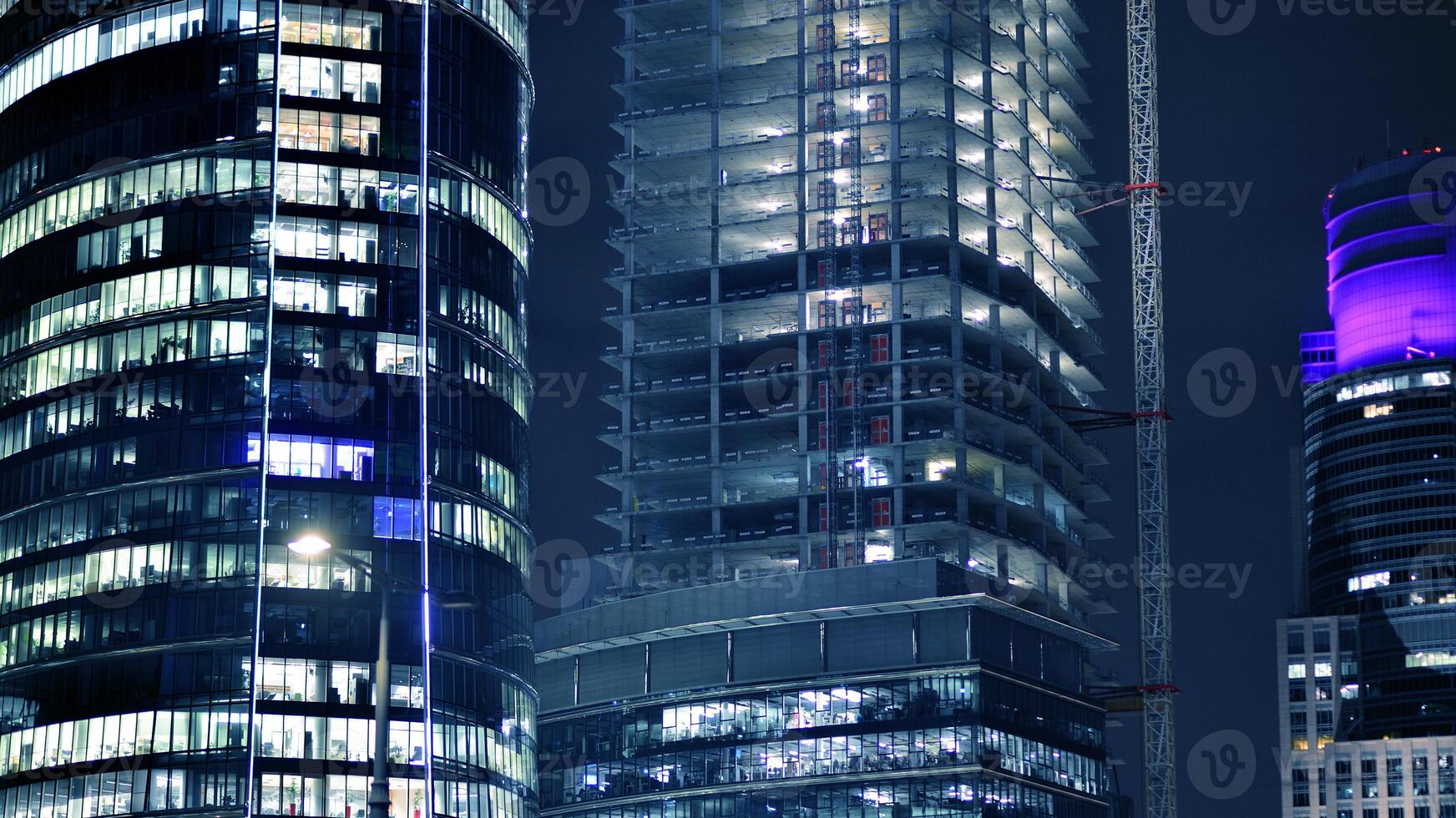 Skyscraper at construction process by night. Modern high-rise building under construction next to glass skyscrapers by night. photo