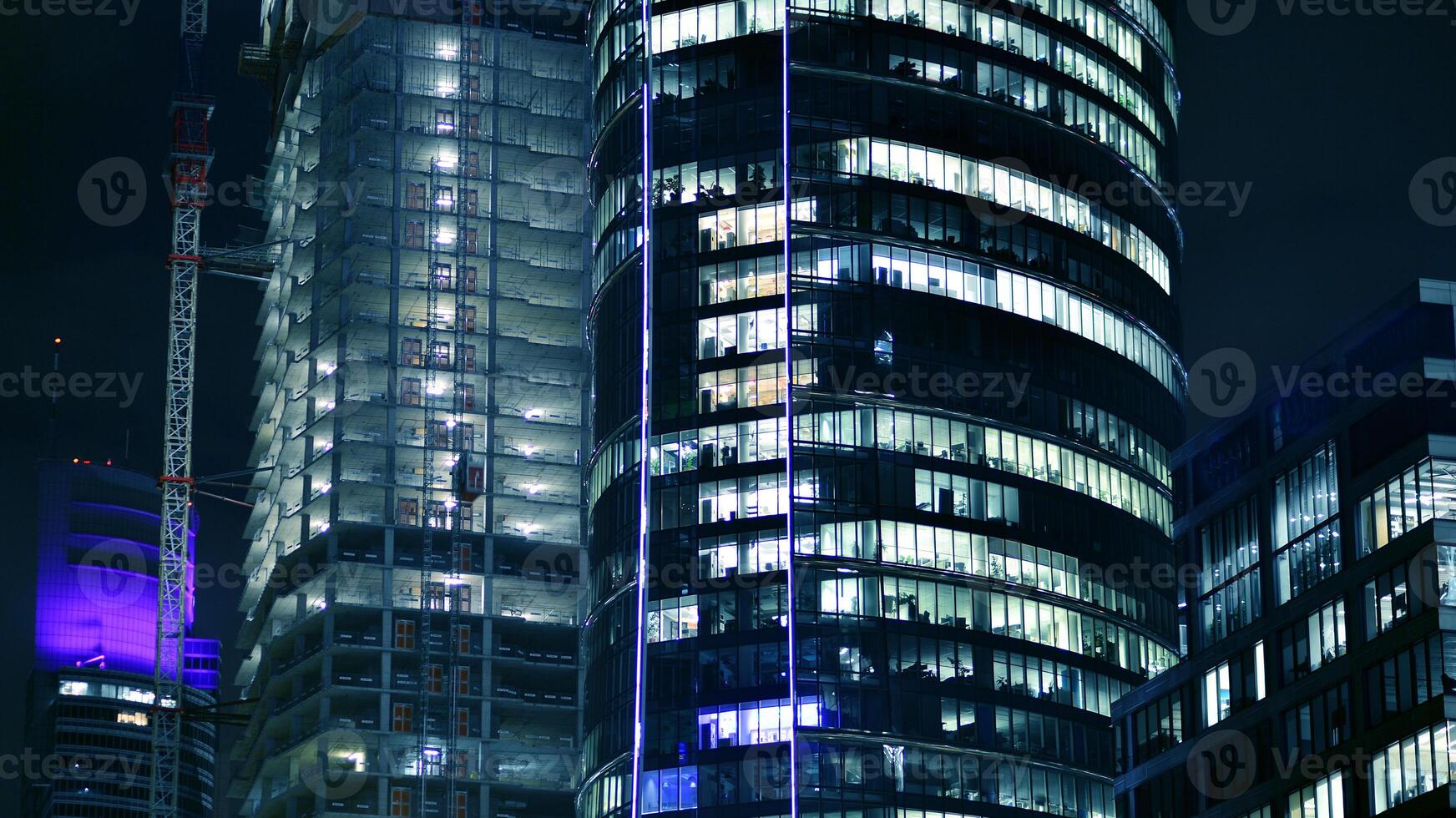 Skyscraper at construction process by night. Modern high-rise building under construction next to glass skyscrapers by night. photo