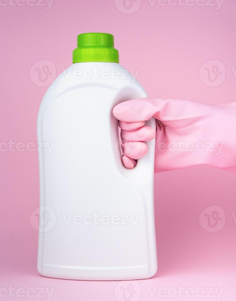 A hand in a pink rubber glove holds a bottle of laundry conditioner, softener bottle, liquid laundry detergent on a pink background. Close-up. Selective focus. photo