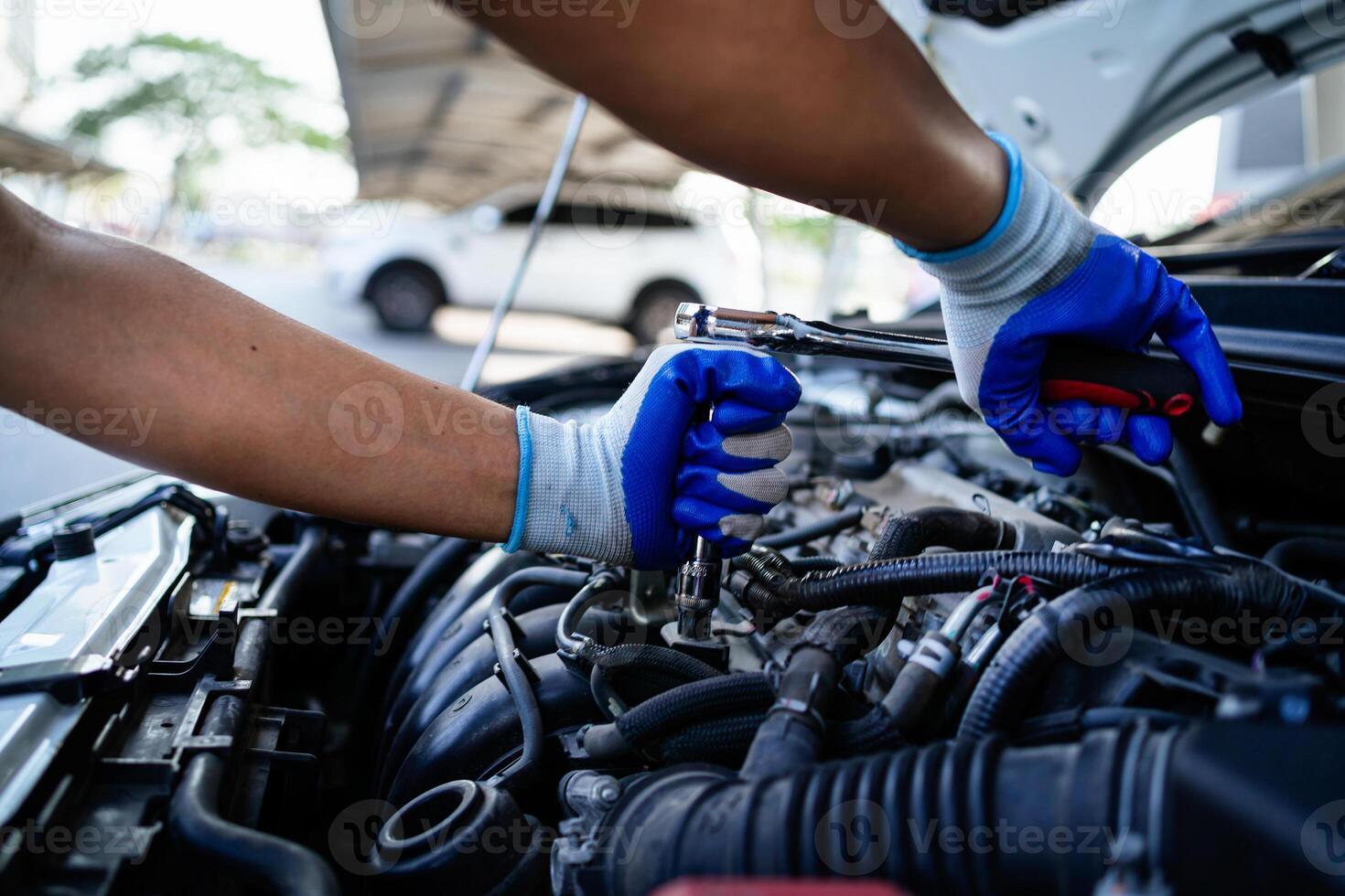 A mechanic is working on a car engine, wearing blue gloves. The car is parked next to a white car photo