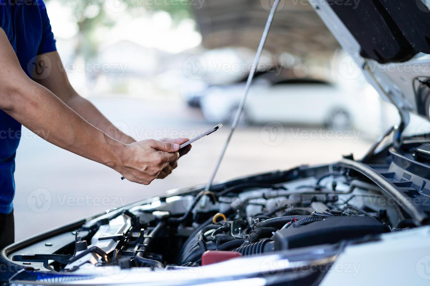 A man in a blue shirt is looking at a tablet while standing next to a car. He is checking the car's engine photo
