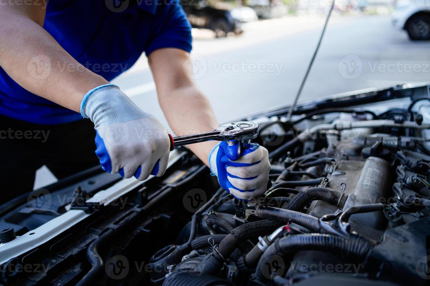 un hombre es trabajando en un coche motor. él es vistiendo guantes y un azul camisa. el coche es estacionado en el lado de el la carretera foto