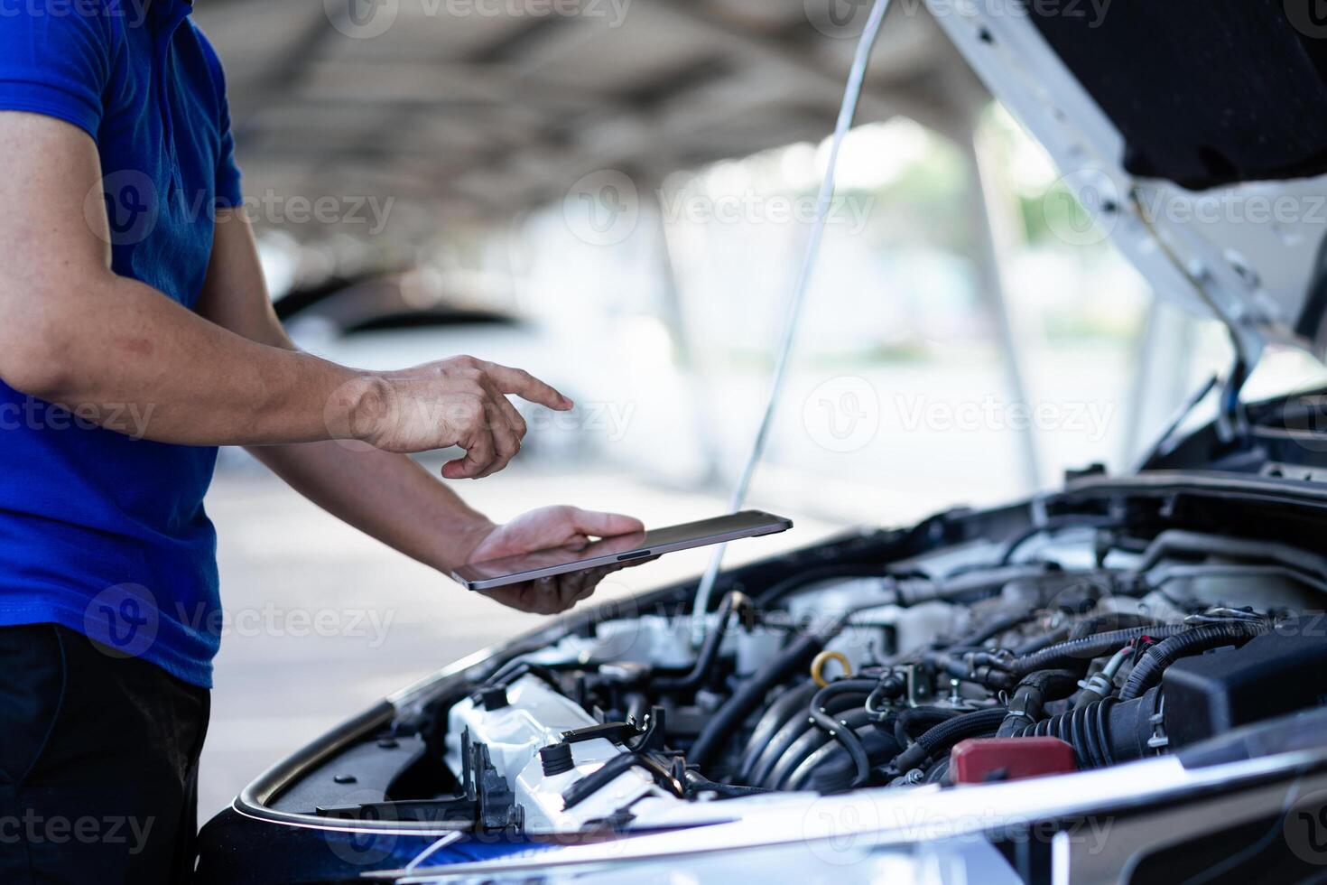 A man in a blue shirt is looking at a tablet while standing next to a car. He is checking the car's engine photo