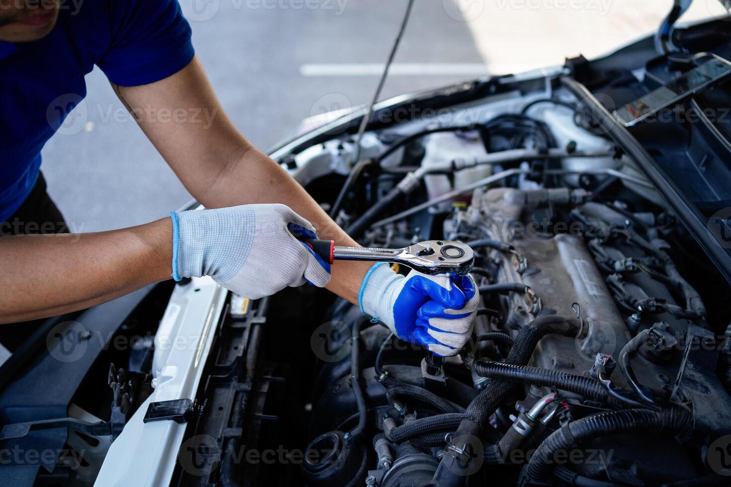A man is working on a car engine. He is wearing blue gloves and a blue shirt. The car is silver and has a hood up photo
