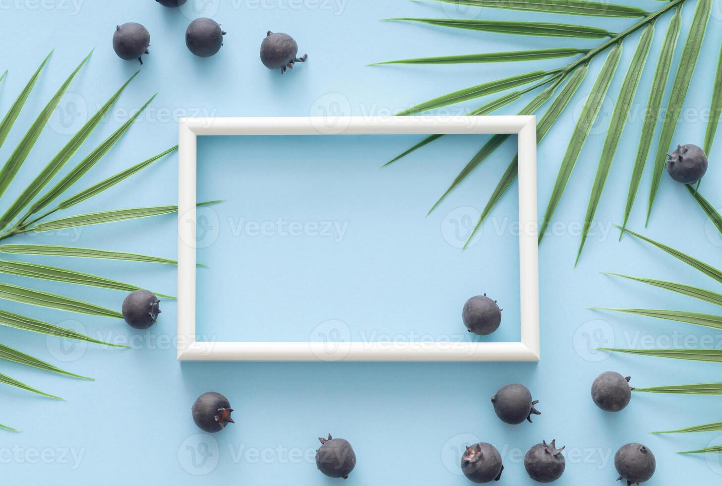 Blueberries and tropical palm leaves with white frame copy space on light blue background. Creative minimal healthy food concept. Trendy summer layout with berry fruit and green leaves. Flat lay. photo
