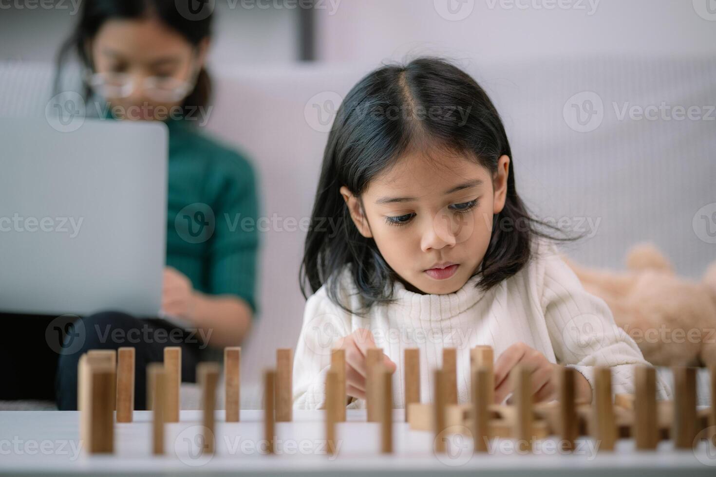 A young girl is playing with wooden blocks while her mother watches photo
