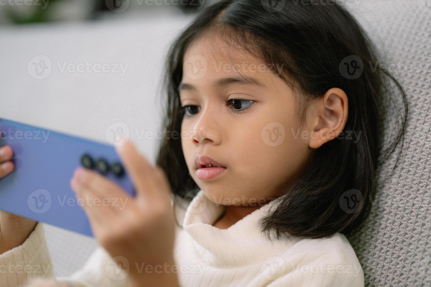 un joven niña es sentado en un sofá y jugando un vídeo juego en un célula teléfono foto