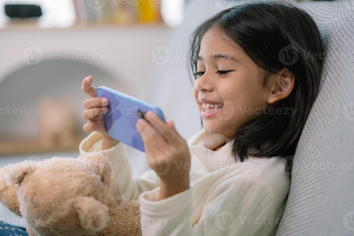 un joven niña es sentado en un sofá y jugando un vídeo juego en su teléfono foto