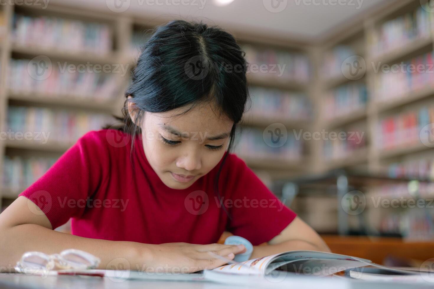 un niña es sentado a un mesa en un biblioteca, leyendo un libro foto