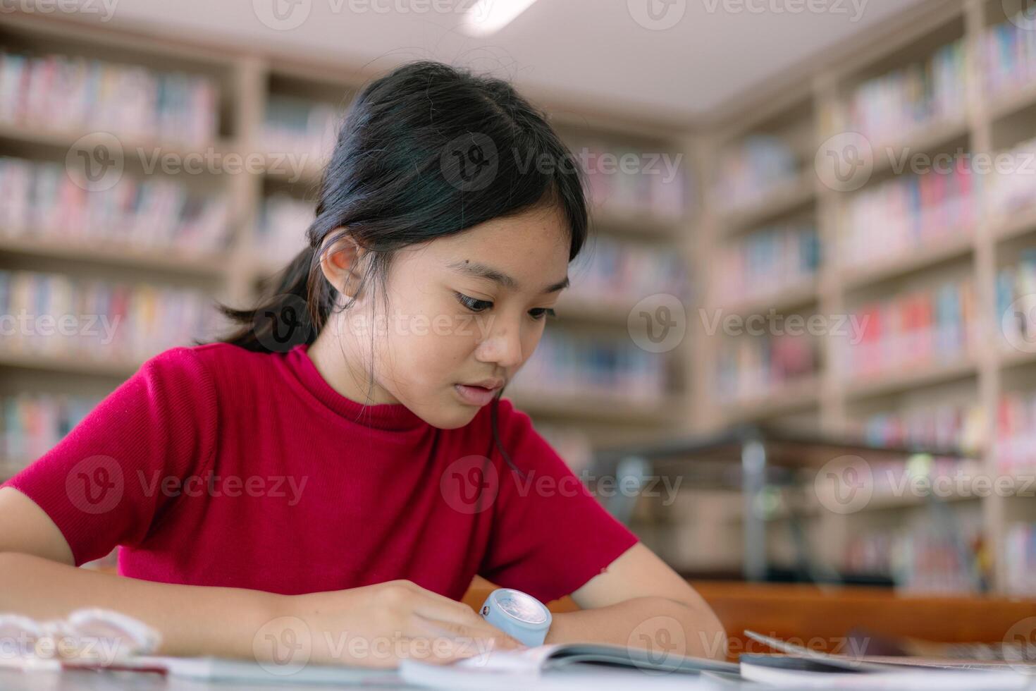 un niña es sentado a un mesa en un biblioteca, leyendo un libro foto