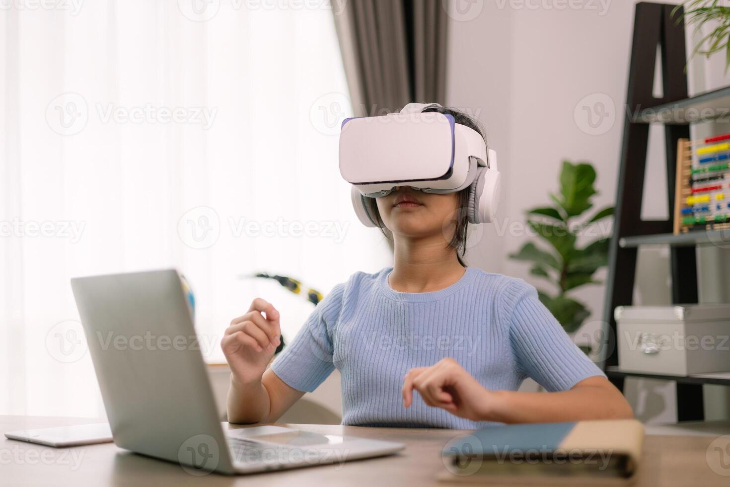 A woman wearing a virtual reality headset is sitting at a desk with a laptop photo