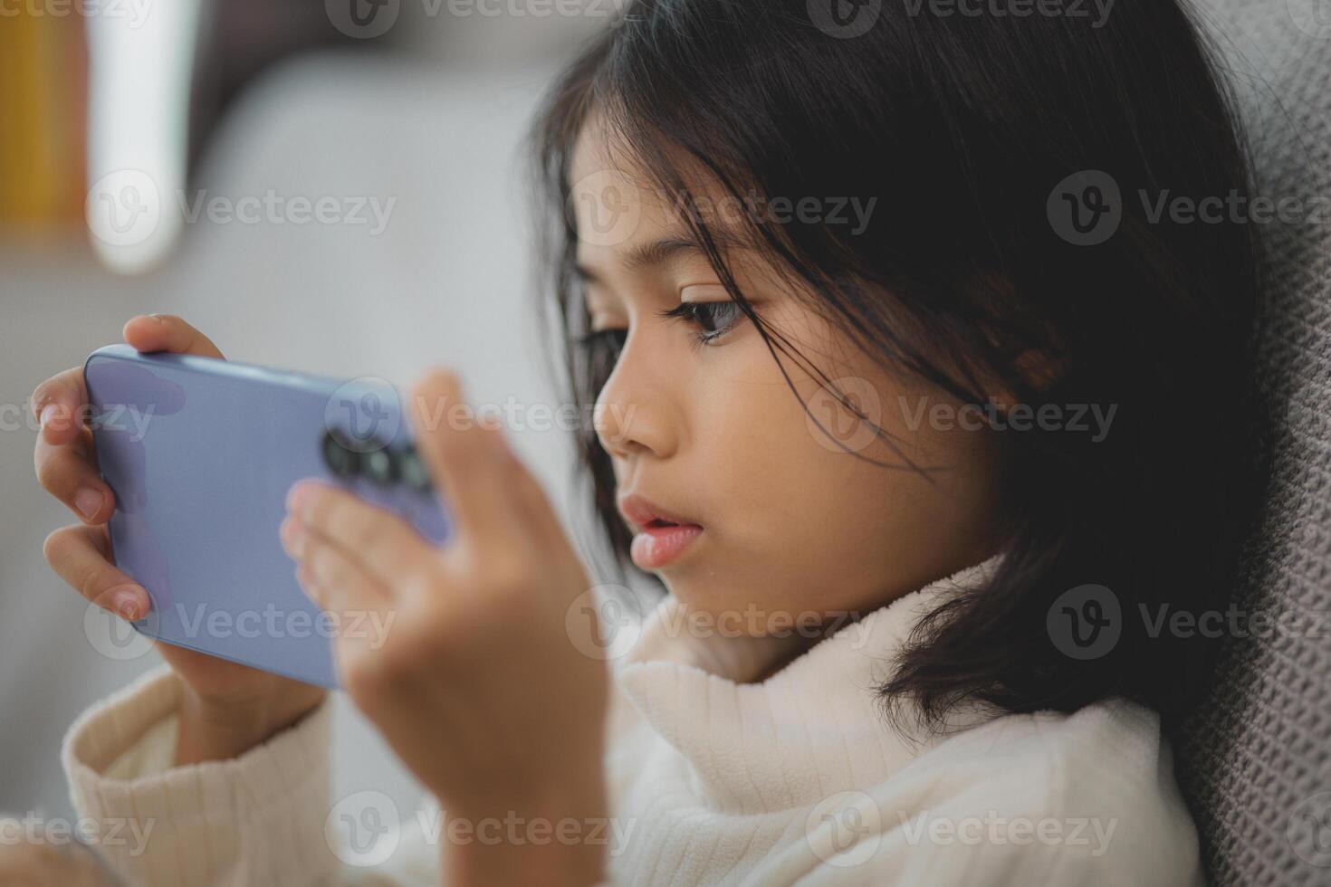 un joven niña es sentado en un sofá y jugando un vídeo juego en su teléfono foto
