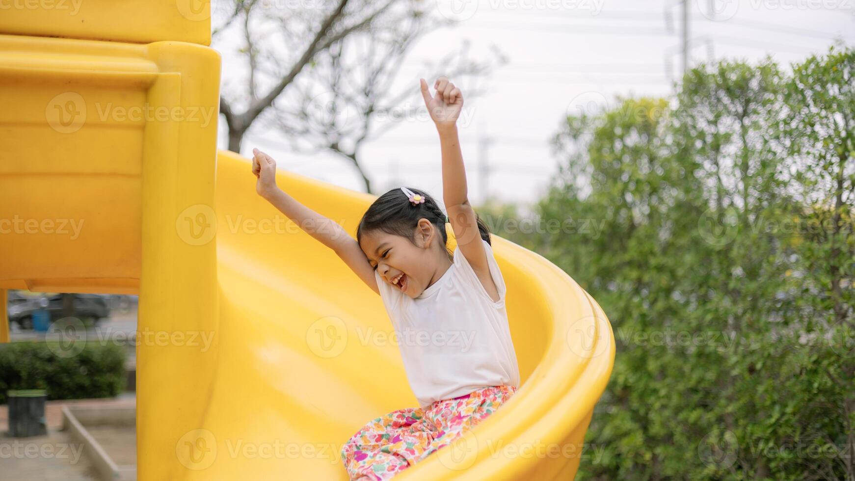 un joven niña es sentado en un amarillo deslizar, sonriente y ondulación foto