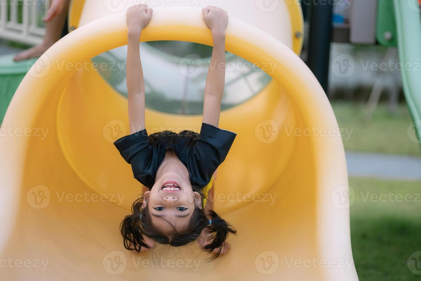 un joven niña es colgando al revés abajo en un amarillo diapositiva foto