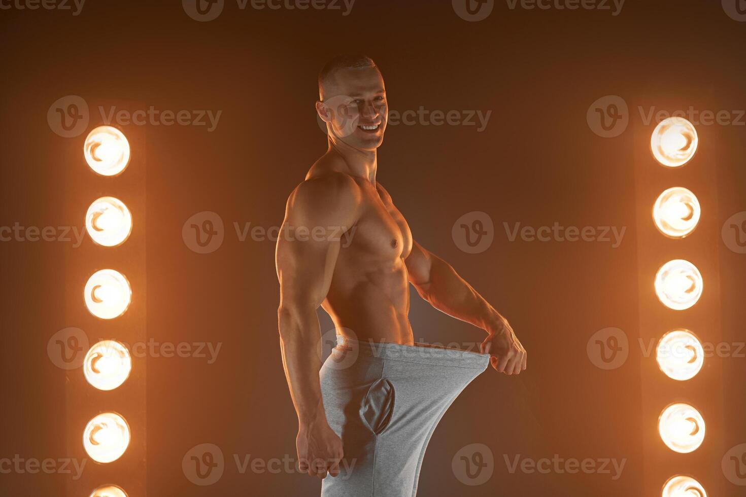 genial masculino tamaño. joven muscular chico tracción espalda su pantalones y demostración pulgar arriba gesto, siendo orgulloso con su genitales, lamparas iluminación en antecedentes foto