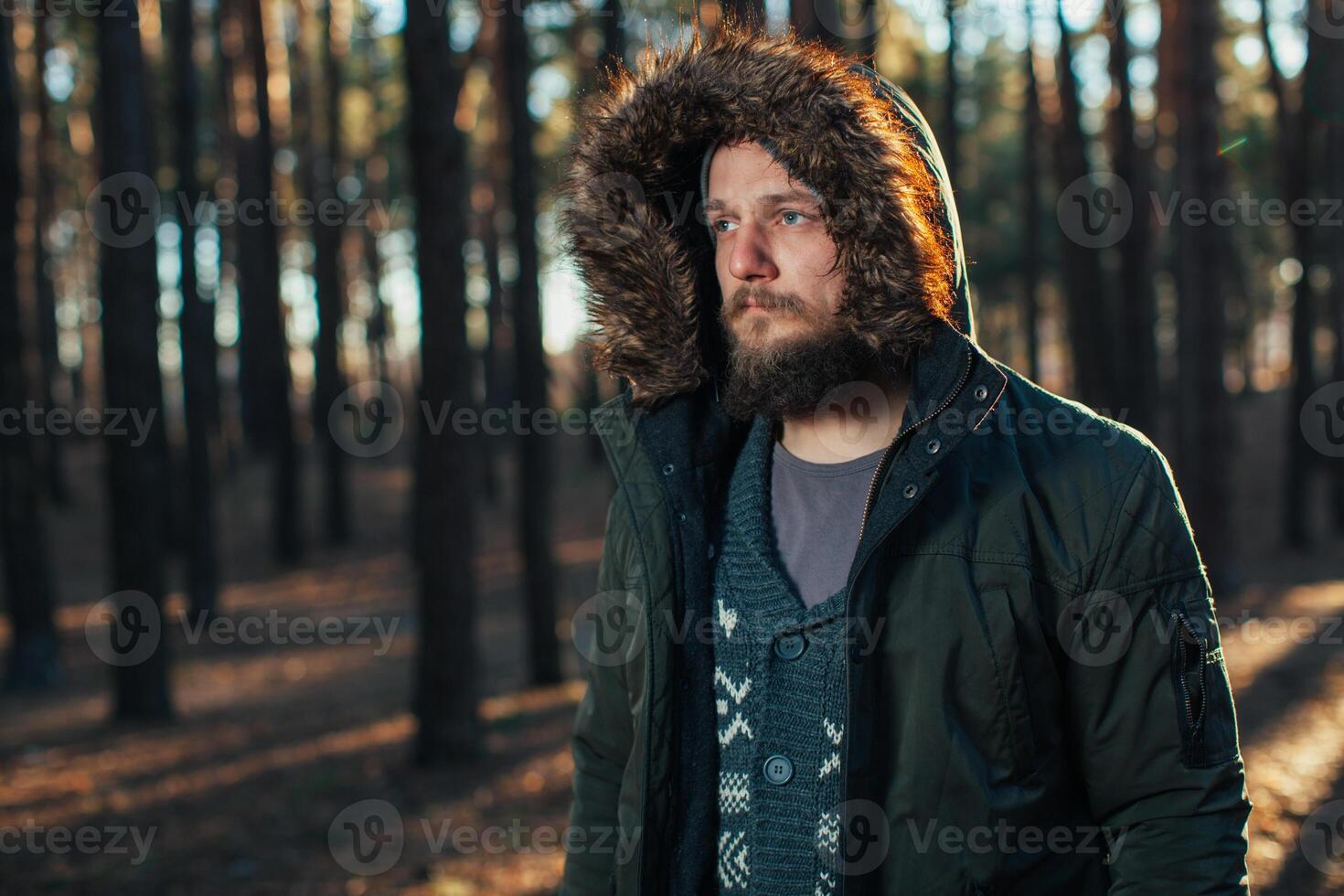 Portrait, close-up of young stylishly serious man with a beard dressed in rgreen winter jacket with a hood and fur on his head stands against the background of pine forest. photo