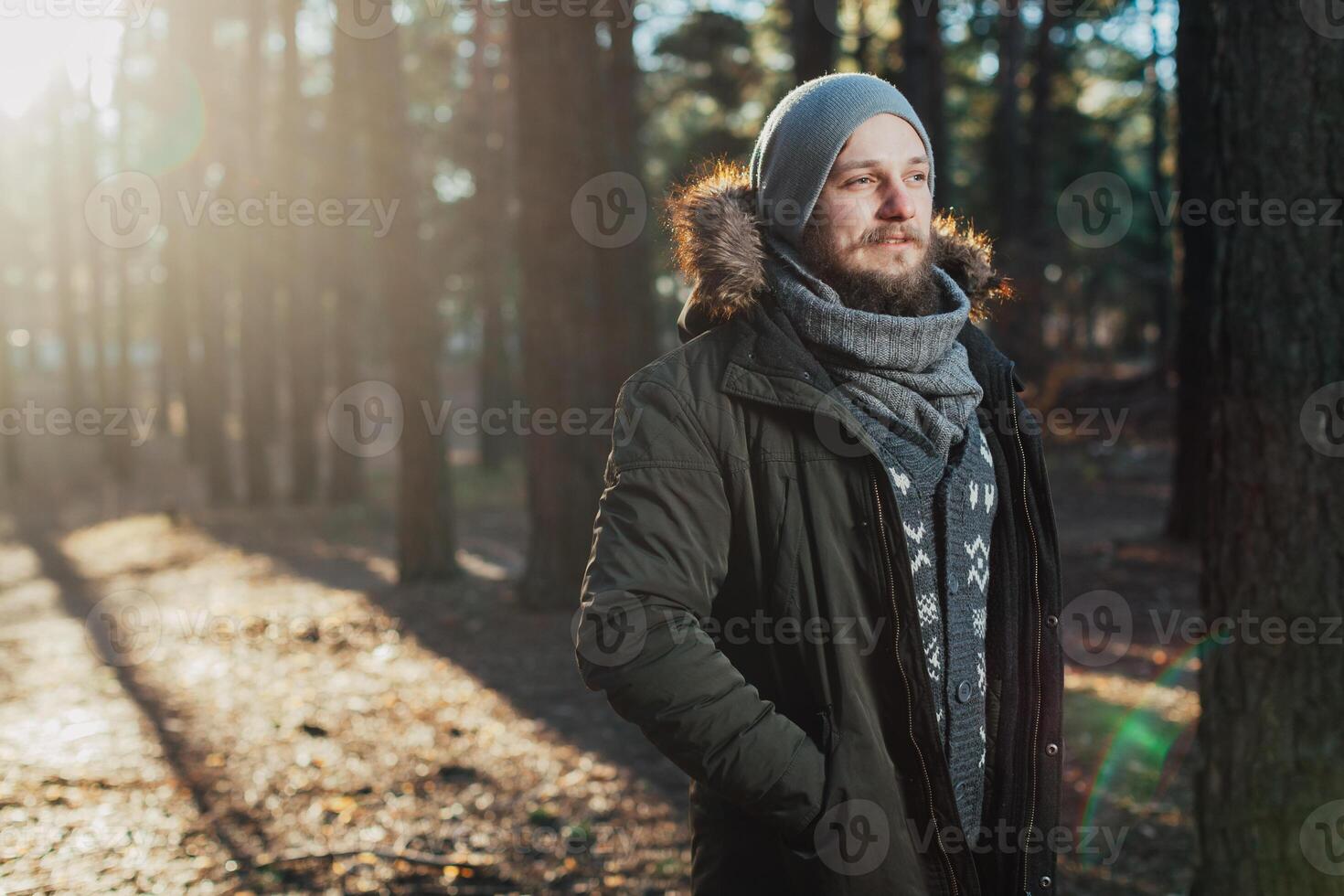 Portrait, close-up of young stylishly serious man with a beard dressed in rgreen winter jacket with a hood and fur on his head stands against the background of pine forest. photo
