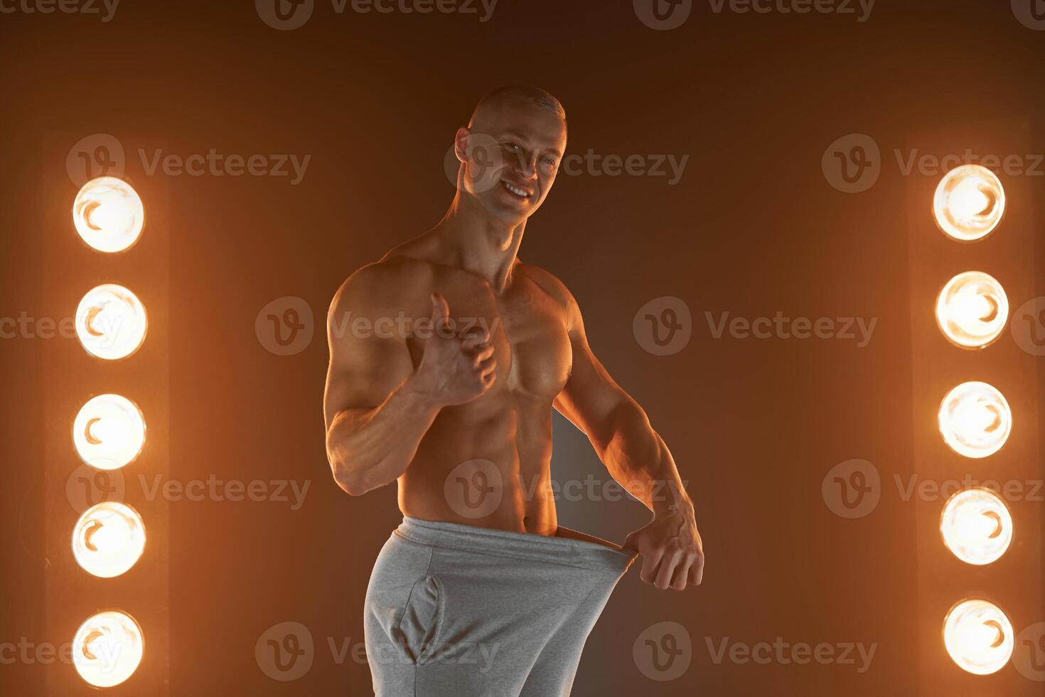 Great male size. Young muscular guy pulling back his pants and showing thumb up gesture, being proud with his genitals, lamps illumination on background photo