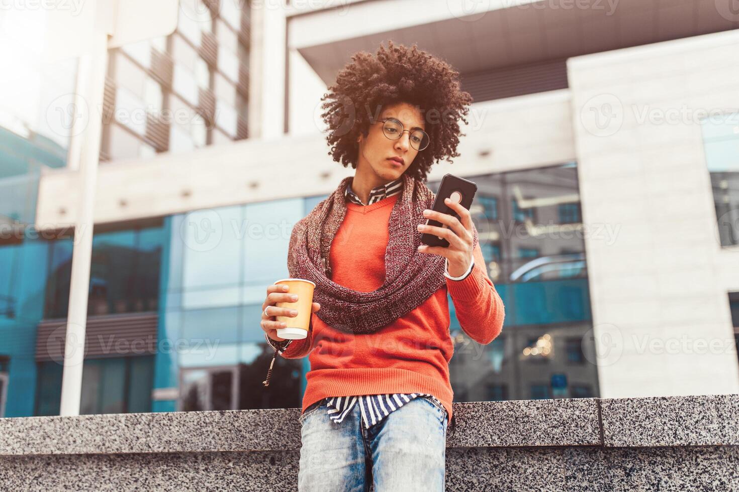 A handsome curly-haired guy of mixed race is dressed in a trendy youth-style scarf and sweater writes a message in a teleone against the background of office buildings. photo