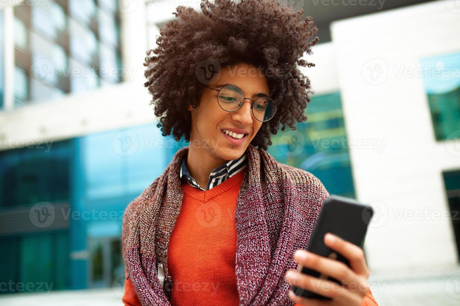 A handsome curly-haired guy of mixed race is dressed in a trendy youth-style scarf and sweater writes a message in a teleone against the background of office buildings. photo