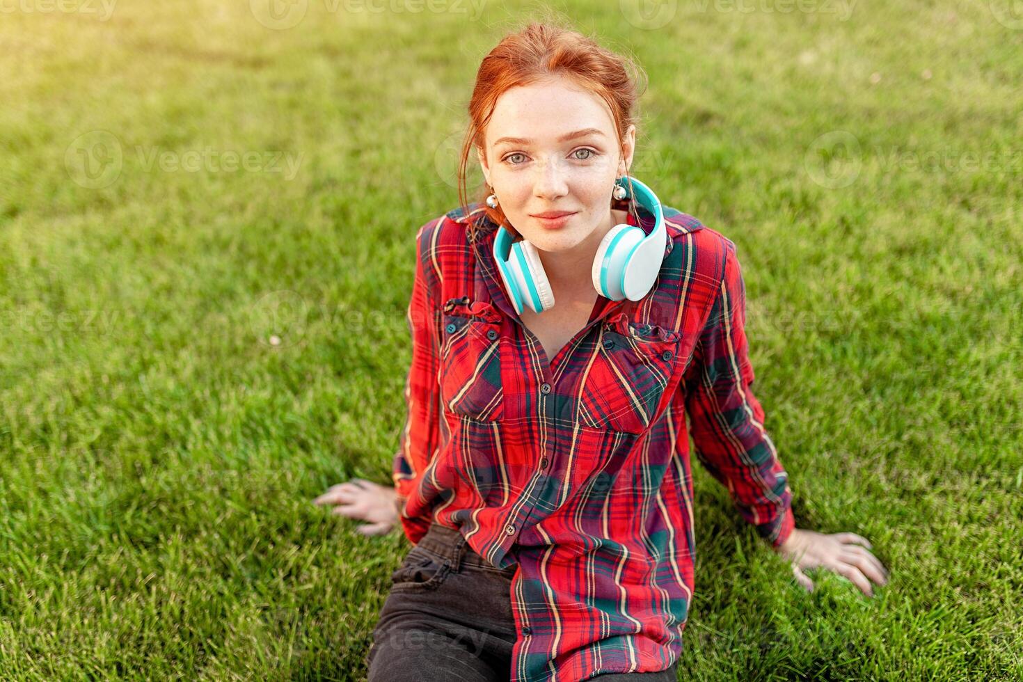 A beautiful red-haired student with freckles is dressed in a red checkered shirt with headphones sitting on the lawn in between the cheba. Student leisure photo